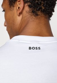T-shirt en coton blanc avec un col côtelé et un logo "BOSS" imprimé au dos, affichant un design épuré et minimaliste.