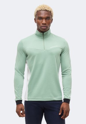 Mann mit kurzen blonden Haaren, der einen hellgrünen Pullover mit Viertel-Reißverschluss, schwarzen Bündchen und marineblaue Hose trägt, steht vor einem einfarbigen Hintergrund.