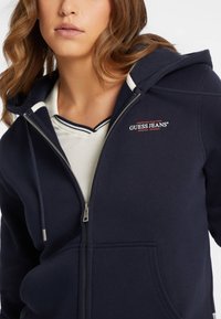 Námornícka mikina na zips s jemnou textúrou a dvoma prednými vreckami. Obsahuje červené vyšívané logo "Guess Jeans" a kapucňu so sťahovacou šnúrkou.