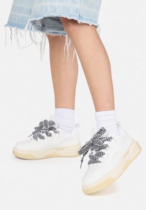 Baskets blanches et épaisses avec des panneaux en maille texturée, des lacets à motifs noir et blanc, et des semelles en caoutchouc beige. Portées avec des chaussettes blanches mi-mollets.
