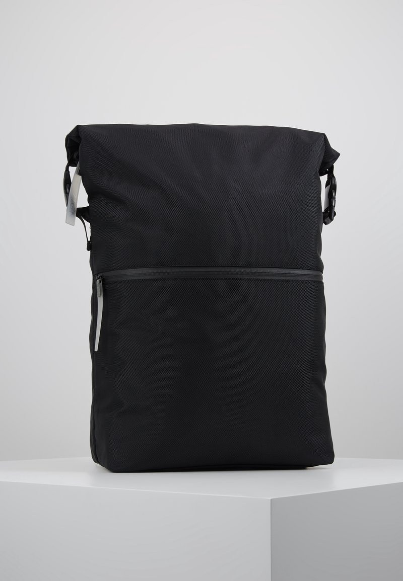 Sac à dos noir à fermeture roulante avec poche zippée à l'avant et boucles latérales, debout sur une surface blanche sur fond gris.