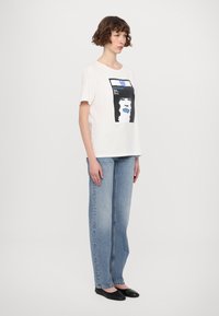 Weißes Baumwoll-T-Shirt mit einem grafischen Design eines Gesichts mit blauen Lippen. Kombiniert mit hellblauen geraden Jeans und schwarzen Ballerinas.