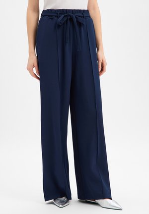 Woman wearing navy wide-leg pants with a tied waistband and silver pointed-toe shoes, standing against a white background.