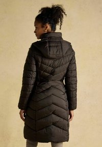 Potažený černý kabát s vzorom chevron, vysokým golierom a pripojenou kapucňou. Má dlhé rukávy a priliehavý pás, zobrazený zozadu.