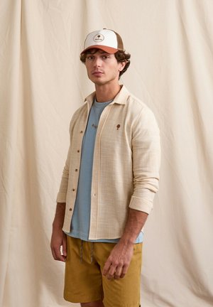 Giovane uomo con una camicia beige abbottonata sopra una t-shirt blu, pantaloncini color senape e un berretto da baseball beige e marrone, davanti a uno sfondo di tessuto chiaro.