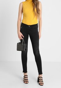 Top sin mangas amarillo con rayas, jeans ajustados negros, bolso negro con tachuelas y sandalias negras con tiras de diseño texturizado.