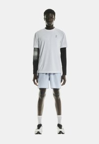 Witte T-shirt met korte mouwen en een zwarte lange mouw eronder, lichtblauwe shorts, zwart-witte sneakers, staande op een effen achtergrond.