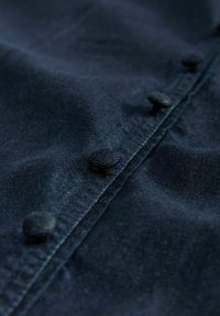 Tissu bleu foncé avec une texture lisse, présentant une rangée de boutons ronds recouverts de tissu le long de la couture. Vue en gros plan du matériau.