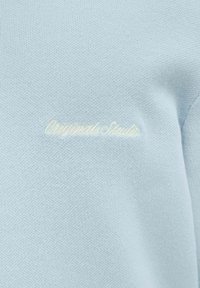 Light blue sweatshirt made of textured fabric, featuring embroidered "Originals Studio" logo in beige. Simple and modern design.