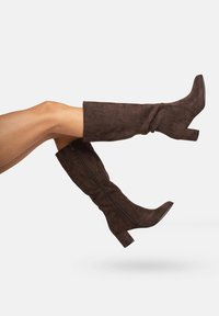 Botas de ante marrón hasta la rodilla con punta cuadrada, tacón grueso y cremallera visible a lo largo del lateral. Textura suave con una silueta ajustada.