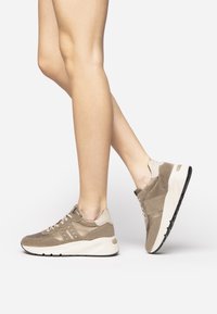 Hellbraune Wildleder-Sportschuhe mit einer klobigen weißen Sohle, versehen mit Schnüren und subtilen Akzenten. Die Seiten- und Rückansicht zeigt das Design.