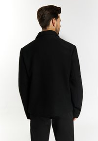 Schwarze Wollmischjacke mit strukturiertem Kragen, oversized Ärmeln und nahtlosem Design, von hinten vor einem weißen Hintergrund betrachtet.