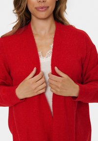 Rode cardigan met een zachte, gestructureerde brei, open voorkant en geribbelde manchetten. Draagt over een lichte, kanten hemdtop.