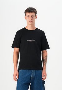 Black cotton t-shirt with short sleeves and rounded neckline. Features a white "JACK & JONES" logo in the center. Worn with blue denim shorts.