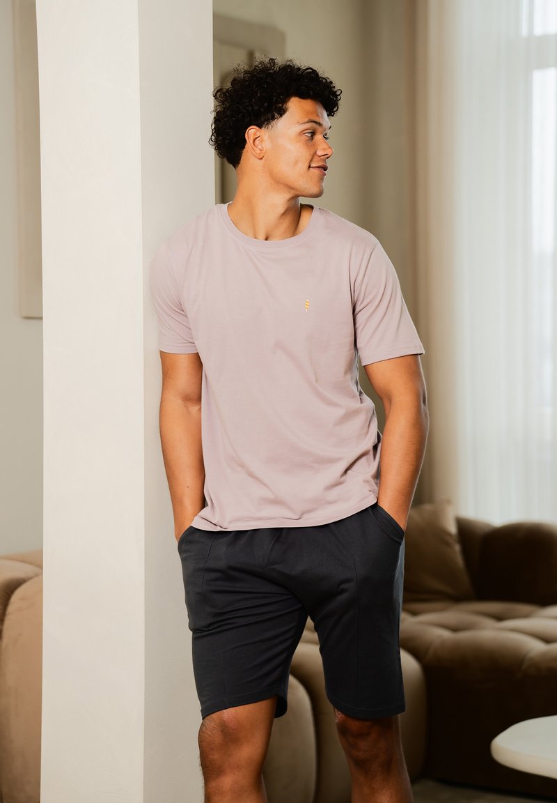 Hellrosanes T-Shirt mit kurzen Ärmeln und einem kleinen gelben Akzent, kombiniert mit dunklen Shorts. Der Stoff wirkt glatt und hat eine lässige Passform.