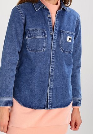 Dunkelblaue Jeanshemd mit Knopfleiste, zwei Brusttaschen, kontrastierenden weißen Nähten und Logopatch auf der Tasche. Über einem rosa Pullover getragen.