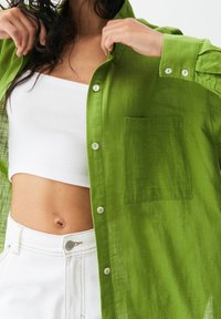 Chemise légère et verte à boutons, avec une finition texturée, dotée d'une poche poitrine et de boutons blancs, portée par-dessus un débardeur blanc à côtes.