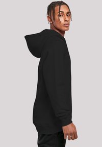 Black hoodie with a kangaroo pocket, adjustable drawstring hood, ribbed cuffs, and a smooth, soft texture. Model stands in profile.