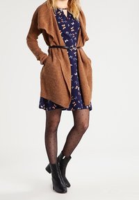 Brun stickad cardigan med öppen front över en marinblå blommig klänning. Svarta prickiga strumpbyxor och klackade anklar stövlar fulländar outfiten.