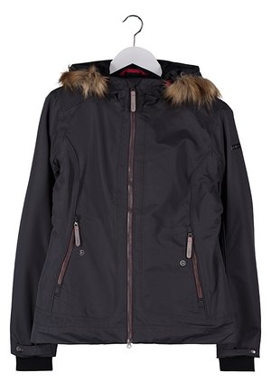Vrouw met een zwarte winterjas met capuchon en bontrand, beige cargobroek en beige wandelschoenen, staand met één hand in de jaszak.