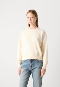 Sweatshirt en coton couleur crème avec une coupe décontractée, des poignets et un ourlet côtelés, arborant un petit logo brodé sur la poitrine. Porté avec un jean bleu.