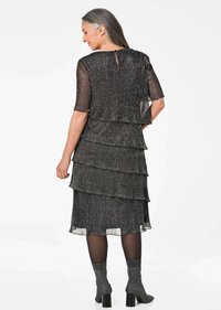 Robe noire à volants avec des manches courtes, présentant un tissu texturé et un détail en trou de serrure à l'arrière. Assortie à des bottes grises à motifs et des collants.