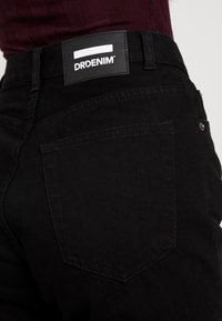 Svart denimjeans med en figurnära design, som har en framträdande märkespatch vid midjan och två bakfickor med kontrasterande sömnad.