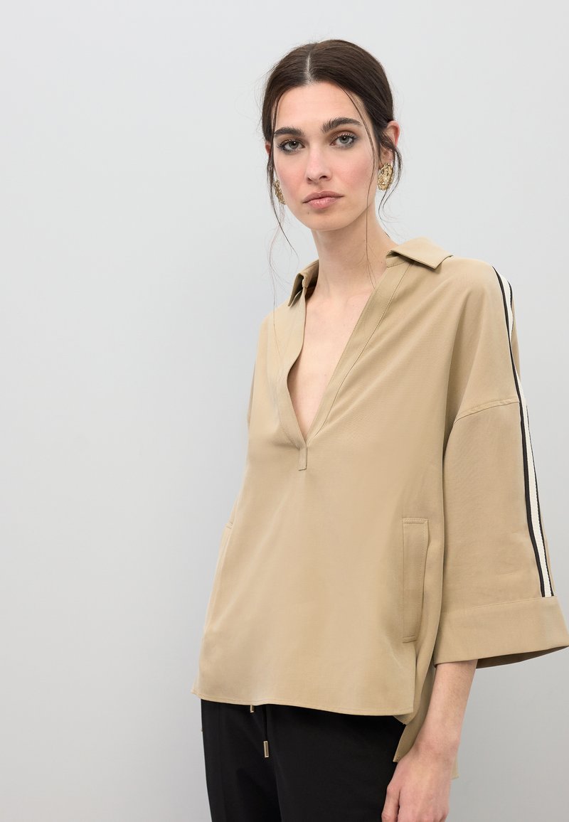 Chemise beige à coupe ample en tissu lisse avec un col en V profond, des manches courtes et des bandes noires sur les côtés. Comprend deux poches latérales.