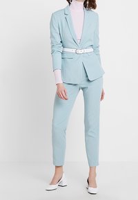 Blazer ajusté bleu clair avec un pantalon assorti, une ceinture blanche cintrée et un col roulé rose. Des chaussures à talons carrés blancs complètent la tenue.