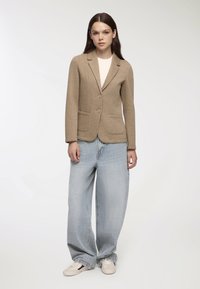 Blazer marrone in lana con design a un bottone, due tasche frontali e revers arrotondati, abbinato a jeans in denim azzurro chiaro e larghi.
