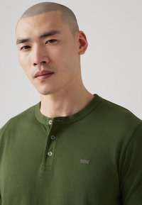 Πράσινη βαμβακερή μπλούζα henley με τρία κουμπιά και μικρό λογότυπο στο στήθος. Λείο υλικό, στρογγυλή λαιμόκοψη και κοντά μανίκια.