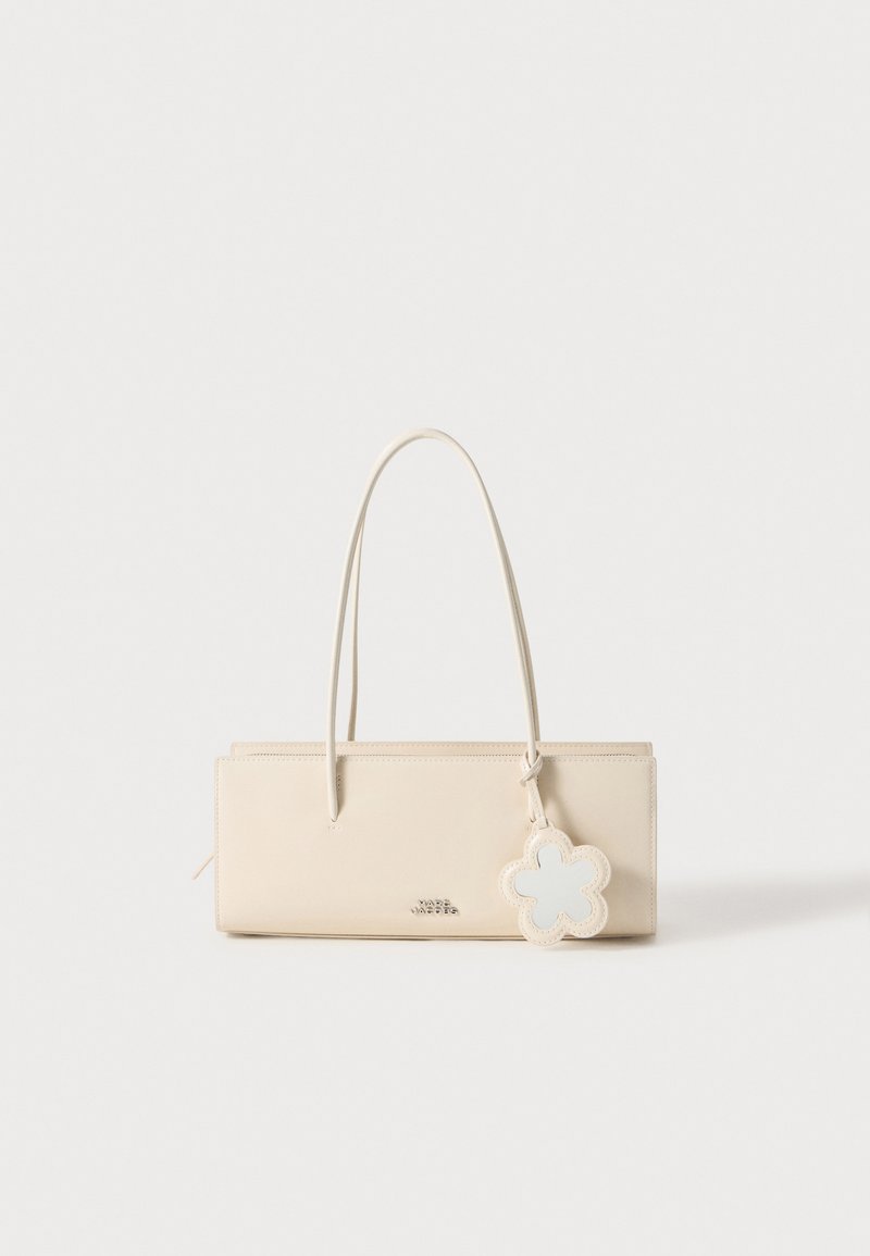 Mala de mão em couro bege com duas alças longas, formato retangular, logotipo em relevo e um charm decorativo em forma de flor azul-clara preso.