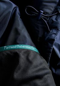 Černá textilní bunda s elastickou páskou se zeleným logem "Portland Outerwear Co." a síťovinovou strukturou na rukávech. Obsahuje černou šňůrku.
