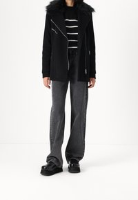 Černý kabát z vlněné směsi s límcem z umělé kožešiny, stříbrné zipy, nošený přes pruhovaný černo-bílý svetr a šedé džíny s širokými nohavicemi. Černé boty na platformě.