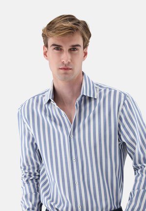 Junger Mann mit hellbraunem Haar trägt ein blau-weiß gestreiftes Hemd mit Knöpfen vor einfarbigem Hintergrund.