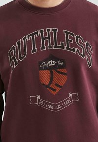 Bordowa bluza z kapturem z napisem "RUTHLESS", tarczą z koroną i banerem z napisem "WYGLĄDAM, JAKBY MNIE TO OBCHODZIŁO" na piersi.