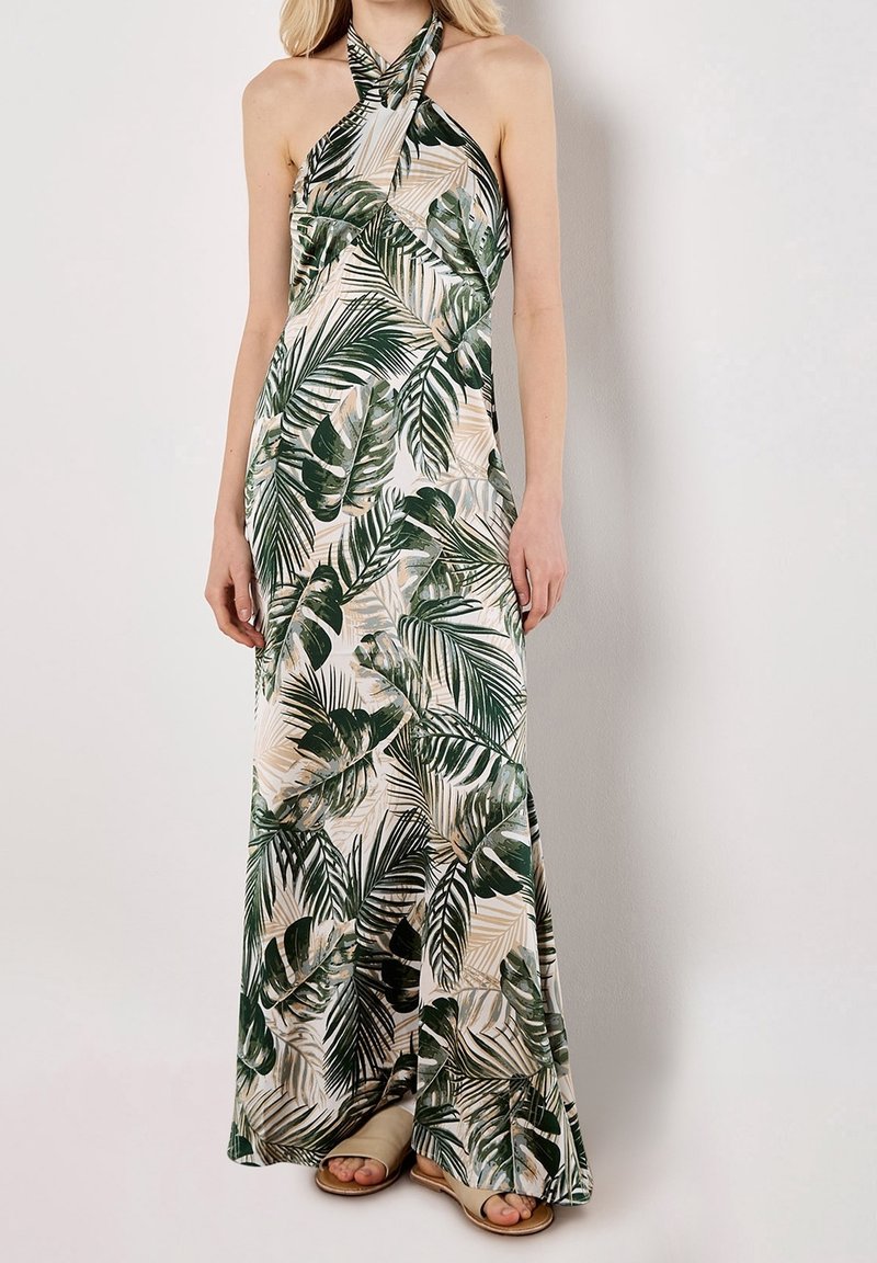 Femme portant une robe maxi sans manches col halter avec un imprimé de feuilles de palmier vertes sur un fond beige clair et des sandales beiges.