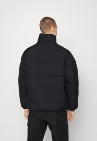 Svart vadderad jacka med hög krage, quiltad design och slät yta, sedd bakifrån. Bärs med mörka cargobyxor.