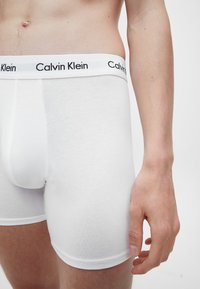Muškarac nosi bijele Calvin Klein bokserice, prikazujući torzo, bok i desnu ruku uz jednostavnu pozadinu.