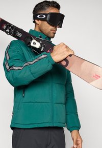 Πράσινη φουσκωτή ζακέτα με μαύρες διακοσμητικές λεπτομέρειες, φορώντας μαύρα γυαλιά σκι, κρατώντας σκι με δεσίματα, σε ουδέτερο φόντο.