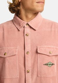 Rosa corduroy-skjorta med krage, som har två bröstfickor och träknappar. En liten broderad logotyp finns på den vänstra fickan.