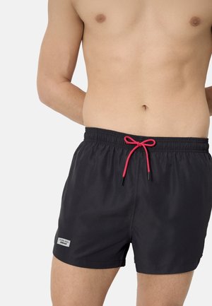 Hombre con pantalones de baño negros con cordón rojo y etiqueta en la pierna izquierda, de pie con las manos detrás de la espalda sobre un fondo claro.