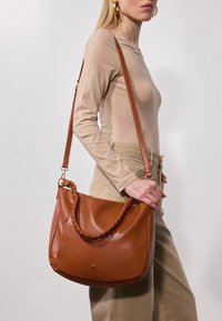 Sac à épaule en cuir marron avec anse tressée, texture lisse, forme semi-structurée, bandoulière réglable et accent de logo discret.