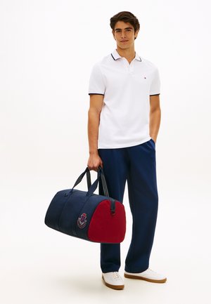 Young man in white polo and navy pants holding a navy and red duffel bag with an embroidered patch, standing against a white background.