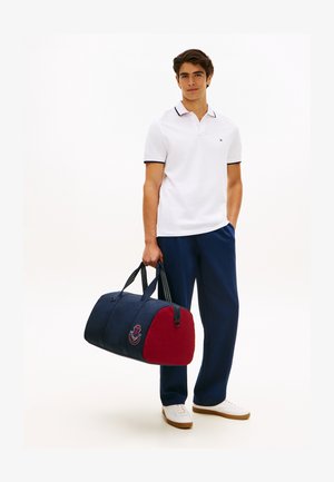 Fiatal férfi fehér pólóban és tengerészkék nadrágban, kezében tengerészkék és piros duffel táskával, amelyen hímzett címke látható, áll fehér háttér előtt.