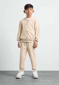 Kreminė užtrauktuku užsegama džemperis su baltu logotipu, derinamas su tokiais pačiais sportiniais kelniais. Papildo baltos sportinės kedai su žaliais akcentais.