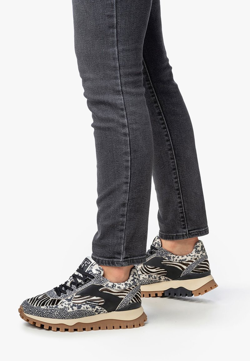 Sneakers con motivo bianco e nero con finitura testurizzata, dotati di suola spessa e design con lacci, indossati con jeans grigio scuro.