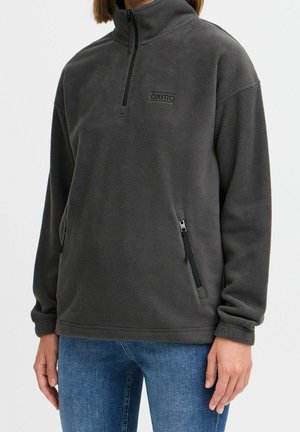 Person iført mørkegrå fleece-pullover med halvlukning, lynlåslommer i siden og blå jeans mod en ensfarvet baggrund.