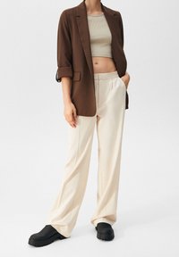 Une personne se tient portant un blazer marron par-dessus un top court beige, associé à un pantalon clair et des chaussures noires, sur un fond uni.