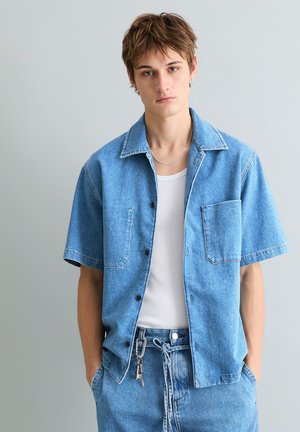 Junge Person, die ein hellblaues Jeanshemd mit kurzen Ärmeln über einem weißen Tanktop und passende Jeanshose mit Schlüsselanhängern trägt.